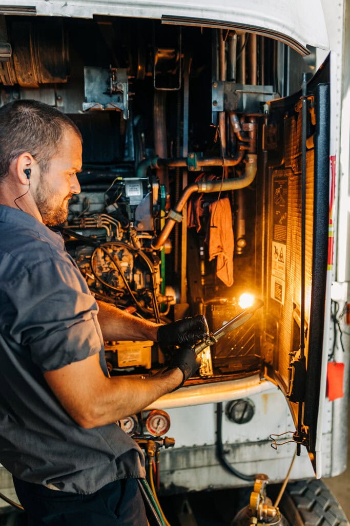 A mechanic, wearing gloves and ear protection, expertly works on a diesel engine with a blowtorch. Surrounded by various engine components and tools, he showcases skills reminiscent of an experienced parts technician while mentoring an entry-level apprentice.