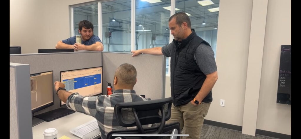 In an office, three men dive into logistics optimization at a computer workstation; two stand while one is seated, gesturing at the screen.