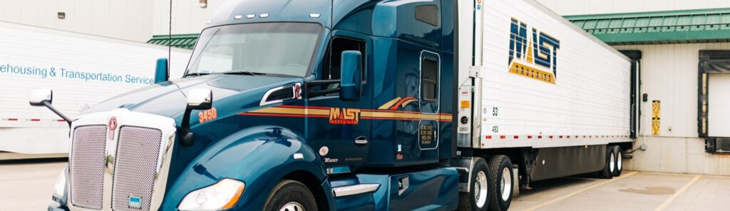 A blue Mast Trucking semi truck is parked in front of a building.